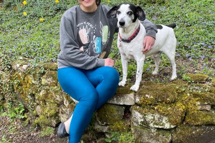 Woman sits with her arm around her black and white dog, on the edge of a garden bed of ivy